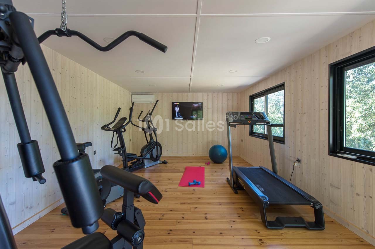 Salle de sport lumineuse avec tapis de course, vélos d'exercice, ballon, tapis de yoga et écran mural.