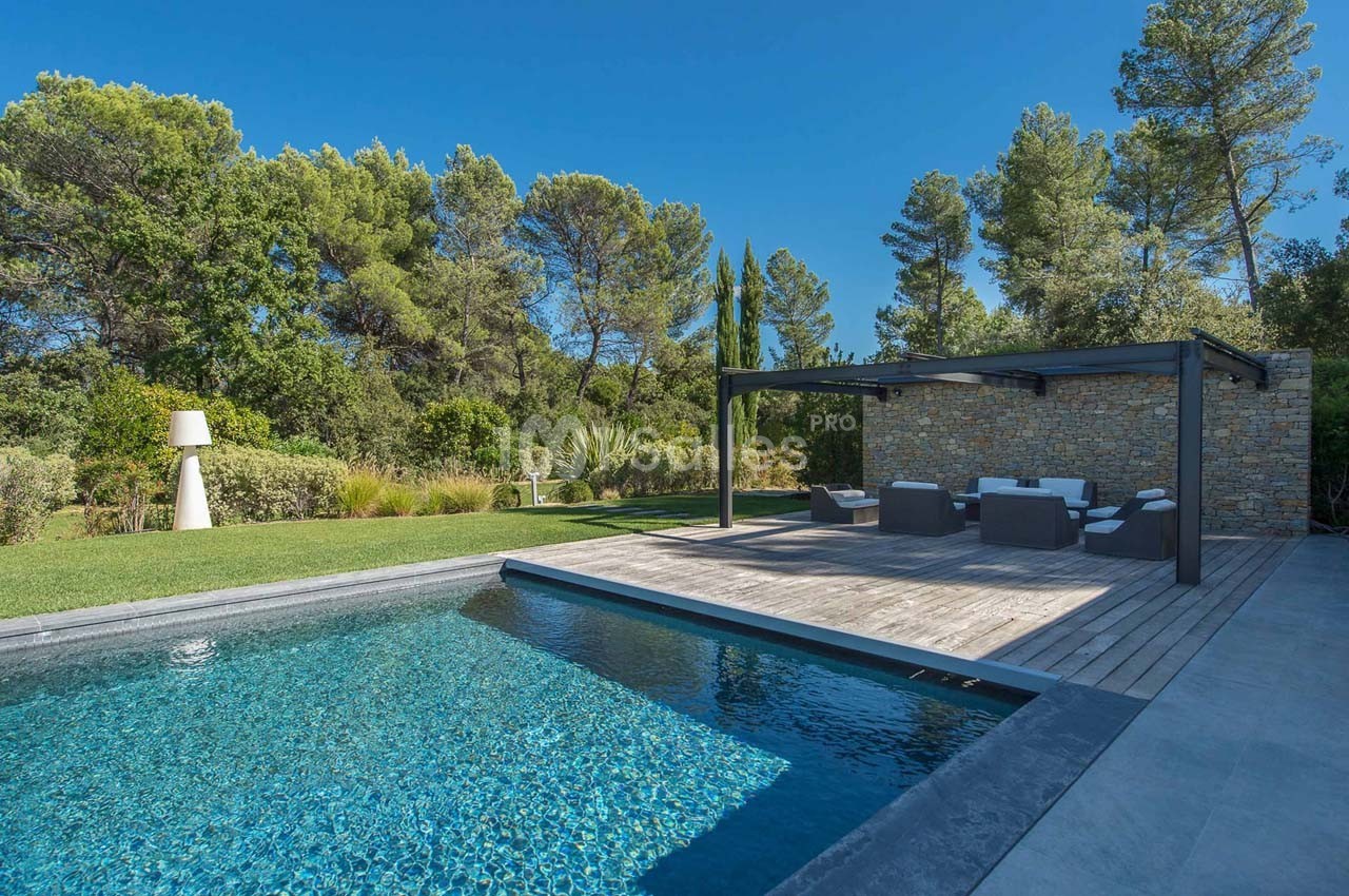 Piscine extérieure entourée de végétation, avec terrasse en bois, salon de jardin et mur en pierre.