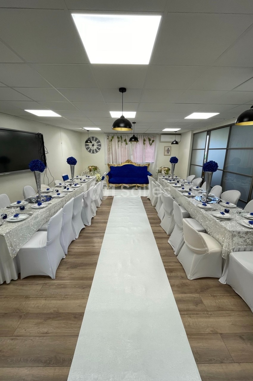 Salle décorée pour un événement, avec tables alignées, nappes blanches, chaises houssées et accents bleus.