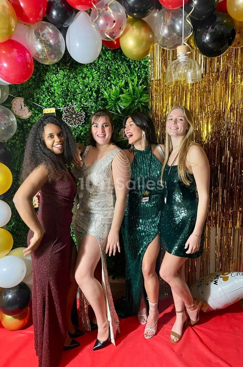 Quatre femmes en robes de soirée posent souriantes devant une décoration festive avec ballons et rideau doré.