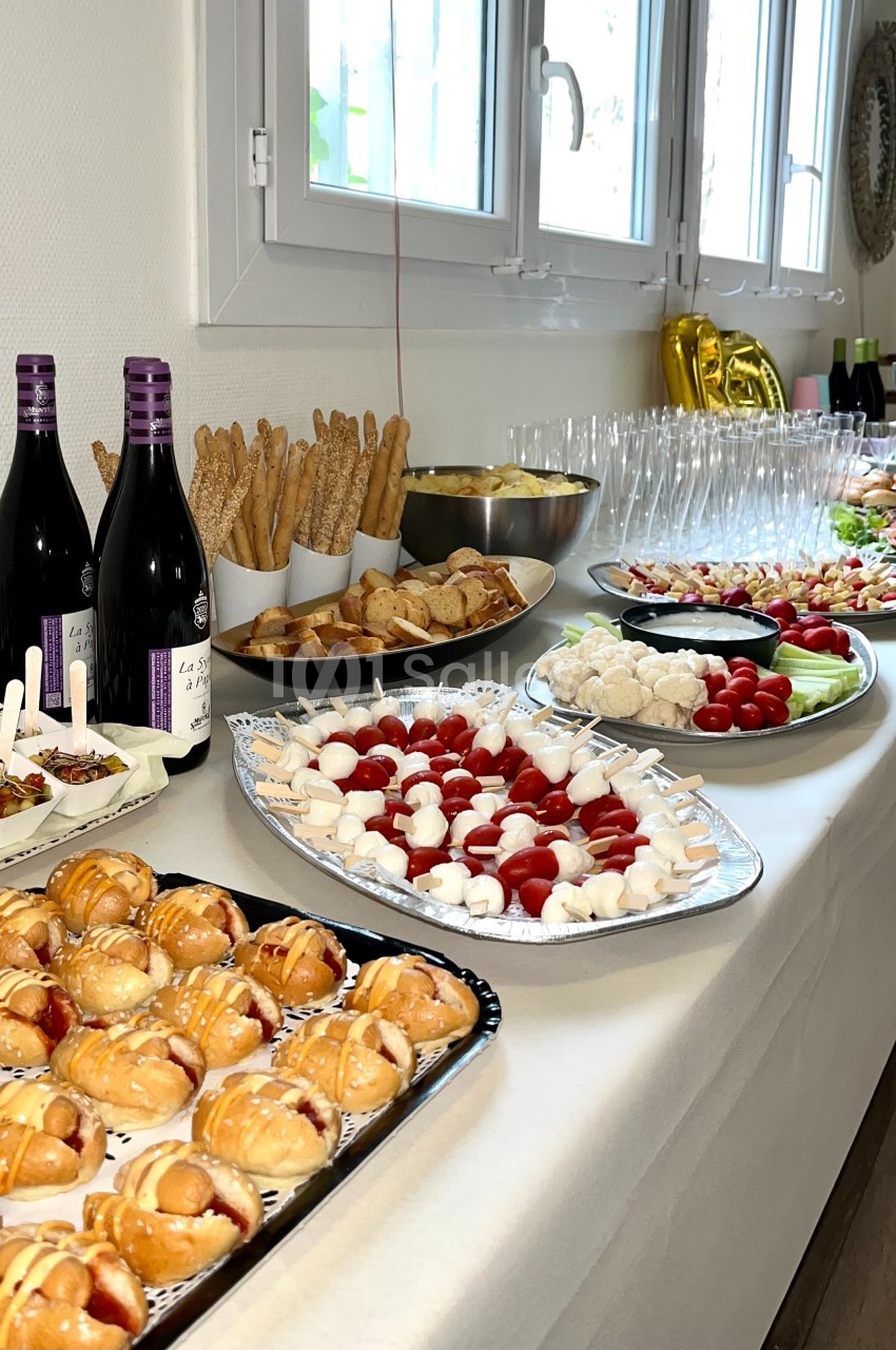 Buffet varié avec viennoiseries, brochettes, crudités, pains, bouteilles de vin et verres disposés sur une table blanche.