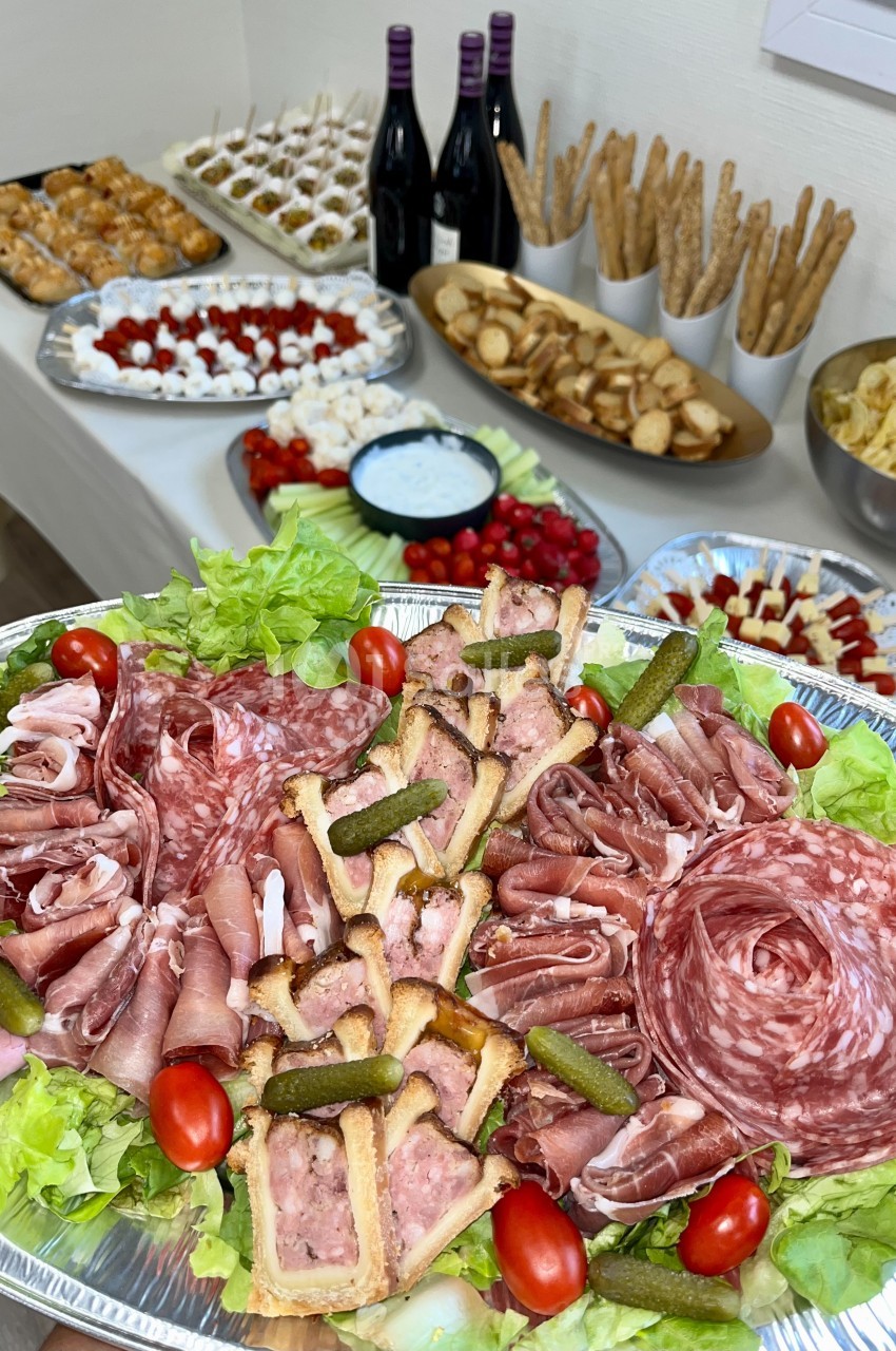 Plateau de charcuterie avec cornichons, tomates cerises et pâtés, entouré d'autres plats et bouteilles de vin en arrière…