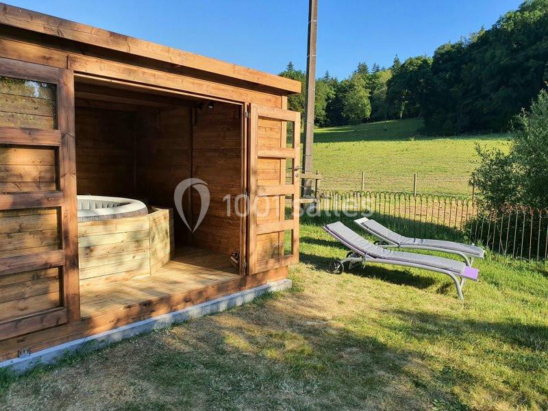 Abri en bois avec un jacuzzi à l'intérieur, deux transats sur une pelouse et paysage verdoyant en arrière-plan.