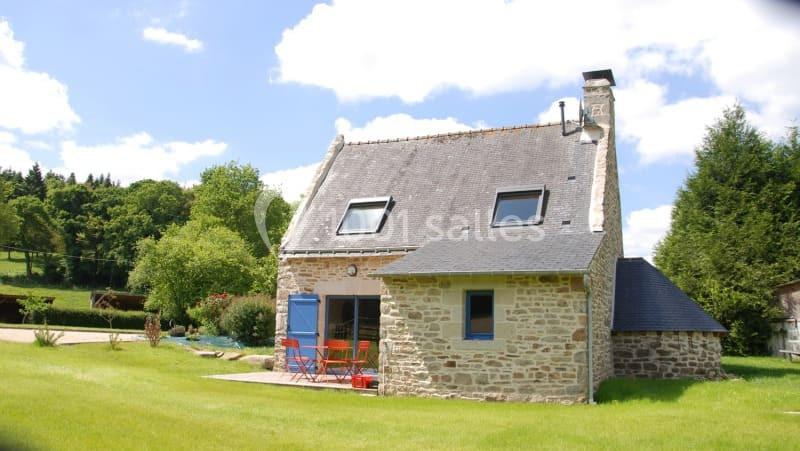 Maison en pierre avec toit en ardoise, entourée de pelouse et d'arbres, sous un ciel bleu avec quelques nuages.