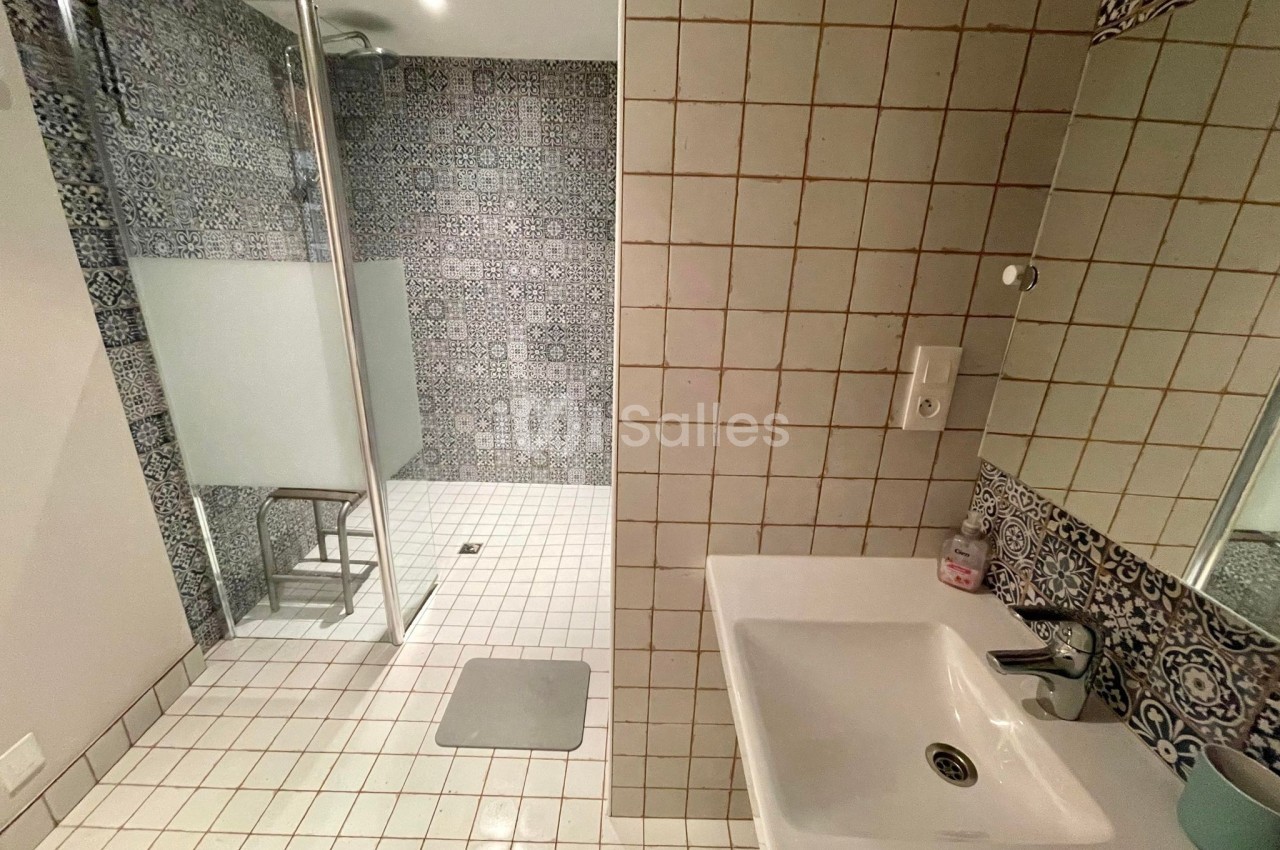 Salle de bain avec douche à l'italienne, carrelage à motifs et mur en carreaux blancs, lavabo avec miroir.