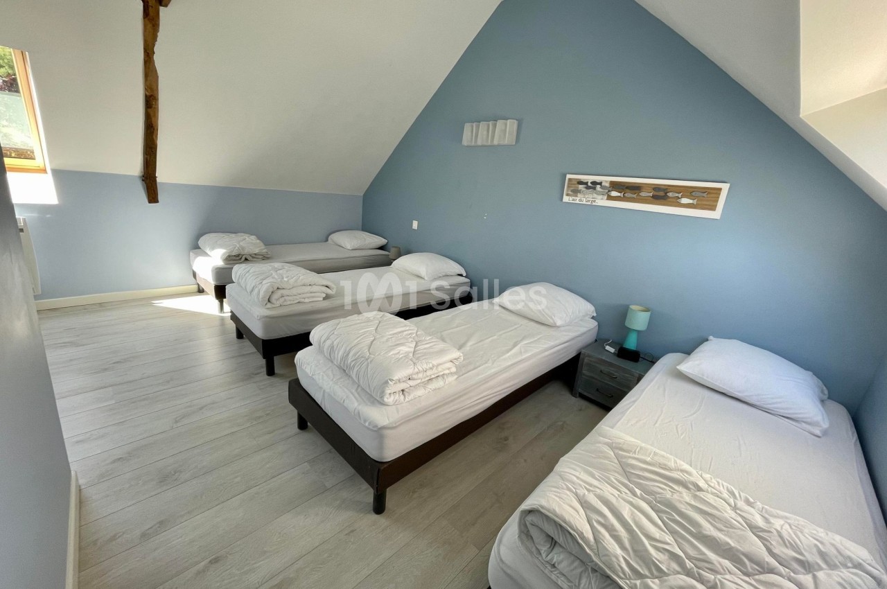 Quatre lits simples alignés dans une chambre mansardée aux murs bleus, avec parquet clair et lumière naturelle.