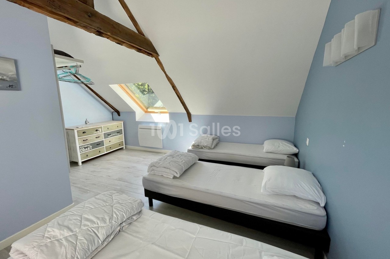 Chambre mansardée avec trois lits simples, murs bleus, poutres apparentes et lumière naturelle provenant d'une fenêtre de…