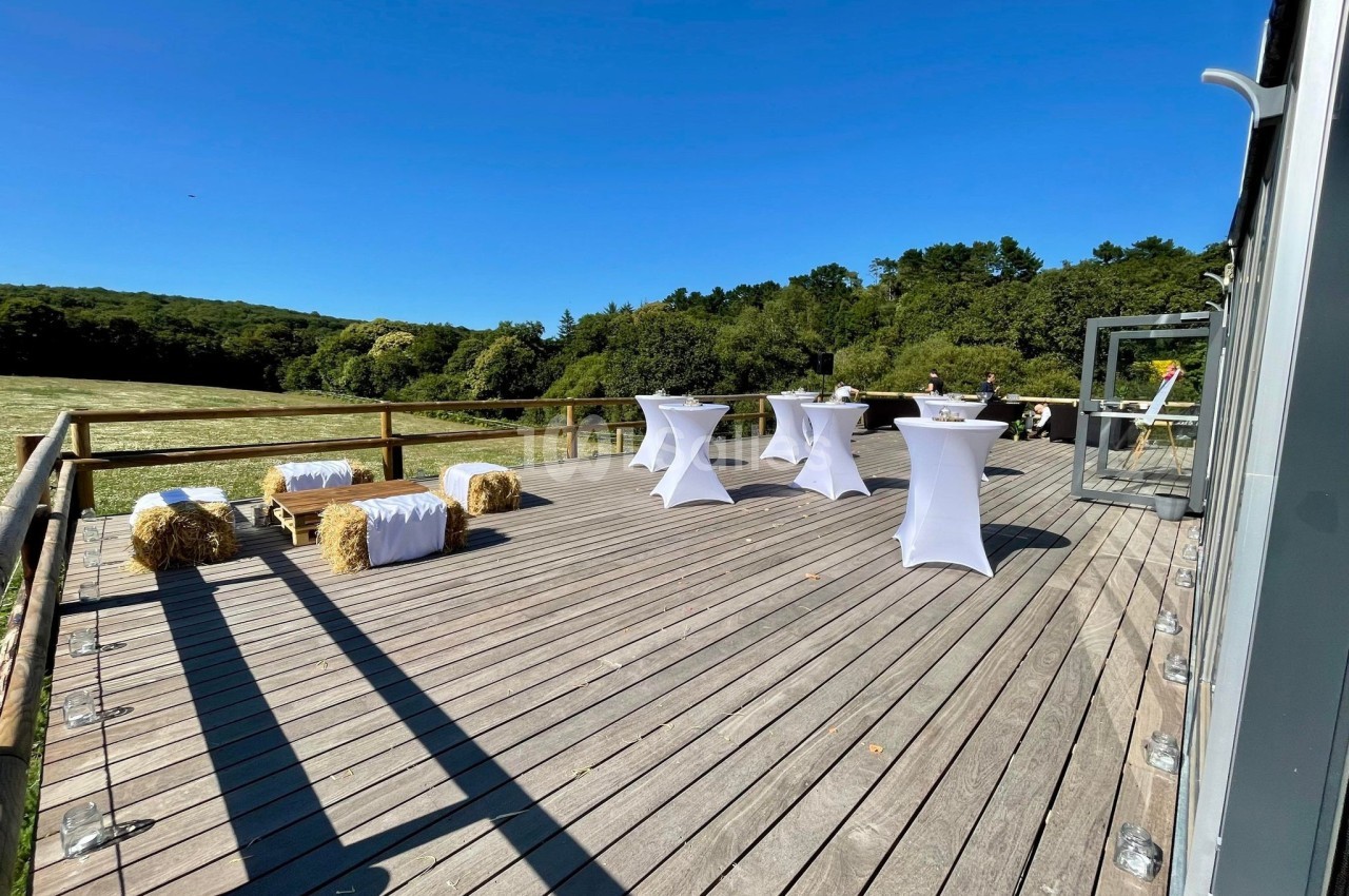 Terrasse en bois avec tables hautes habillées de nappes blanches, bottes de paille et vue sur un paysage verdoyant.