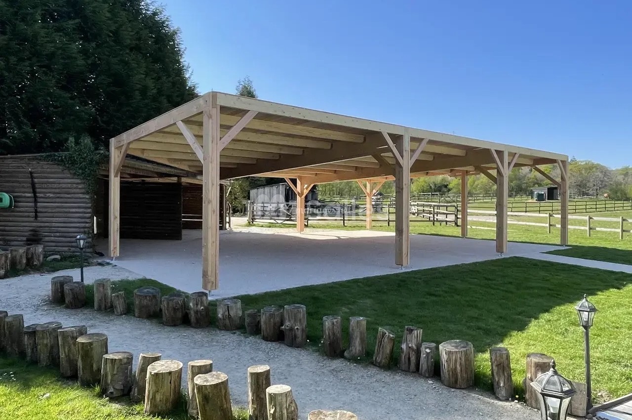Structure en bois ouverte avec toit, située dans un espace extérieur verdoyant, entourée de rondins et de pelouse.
