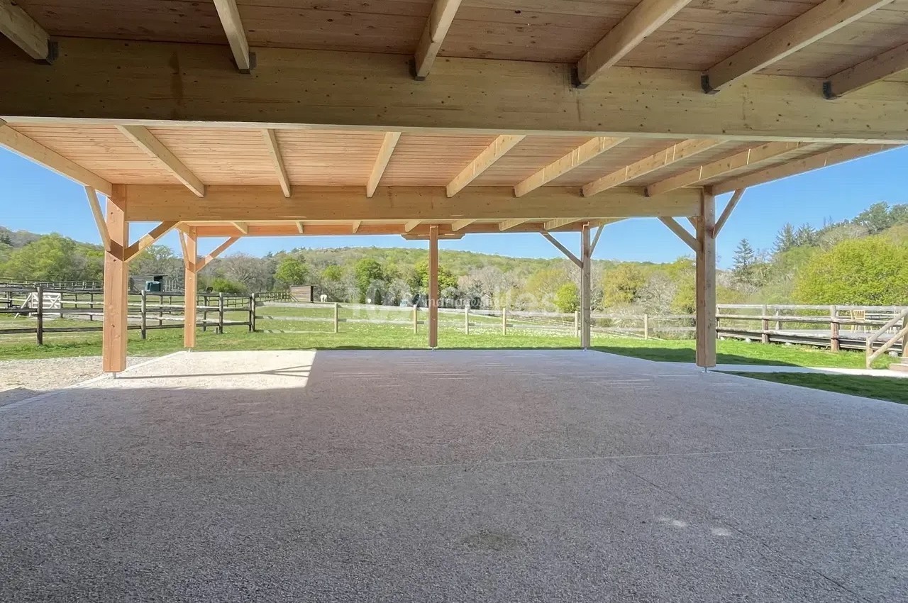 Structure en bois ouverte avec toit, offrant une vue sur un espace extérieur verdoyant et des clôtures en arrière-plan.