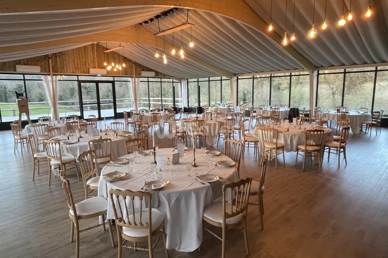 Salle de réception lumineuse avec tables rondes dressées, chaises en bois clair et grandes baies vitrées donnant sur un…