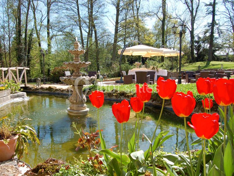 Étang avec une fontaine entouré de tulipes rouges, terrasse aménagée et arbres en arrière-plan.