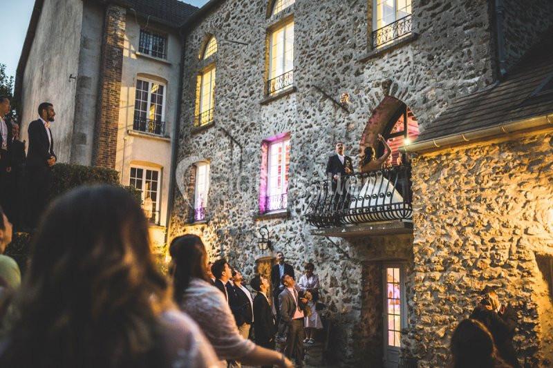 Un groupe de personnes rassemblées devant une maison en pierre éclairée, certaines regardant vers un balcon.