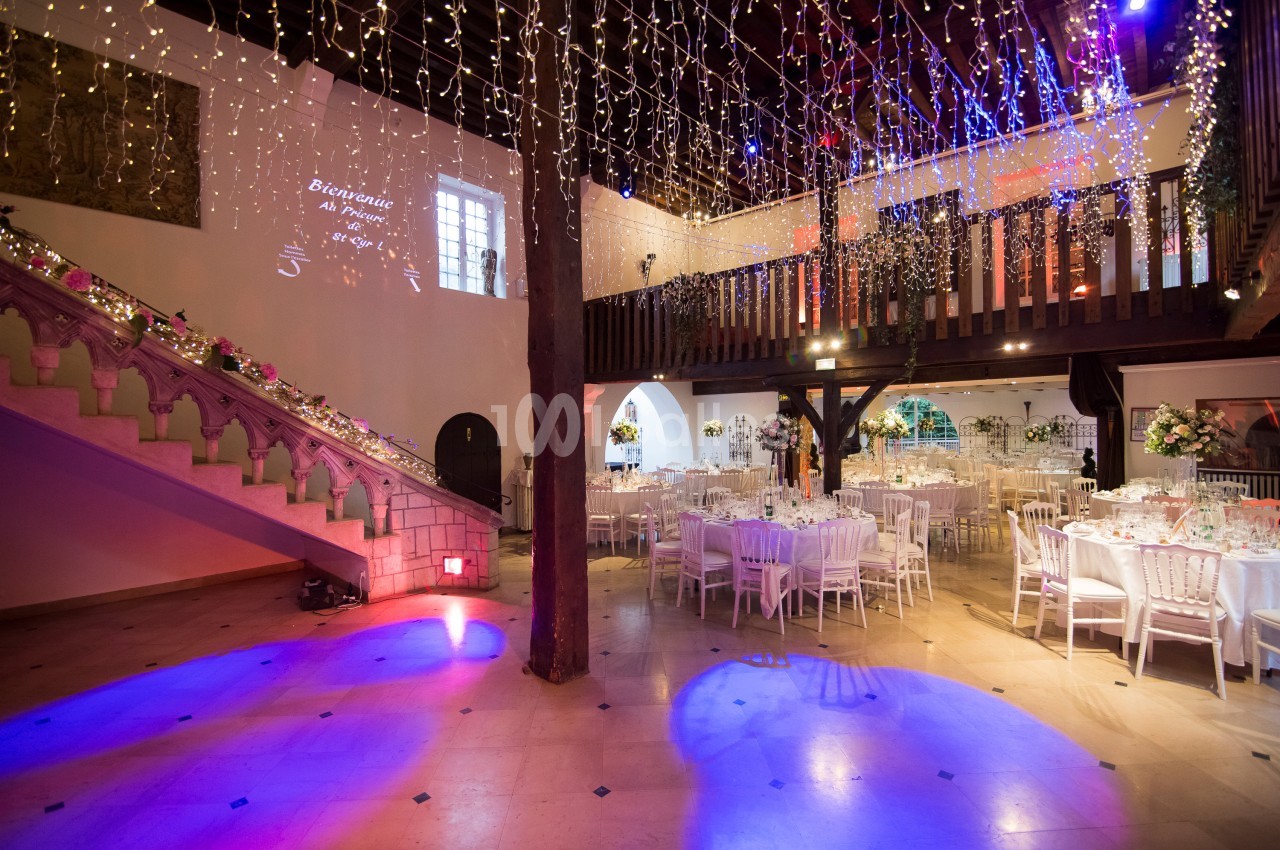 Salle de réception décorée avec des guirlandes lumineuses, tables dressées et escalier orné de fleurs.