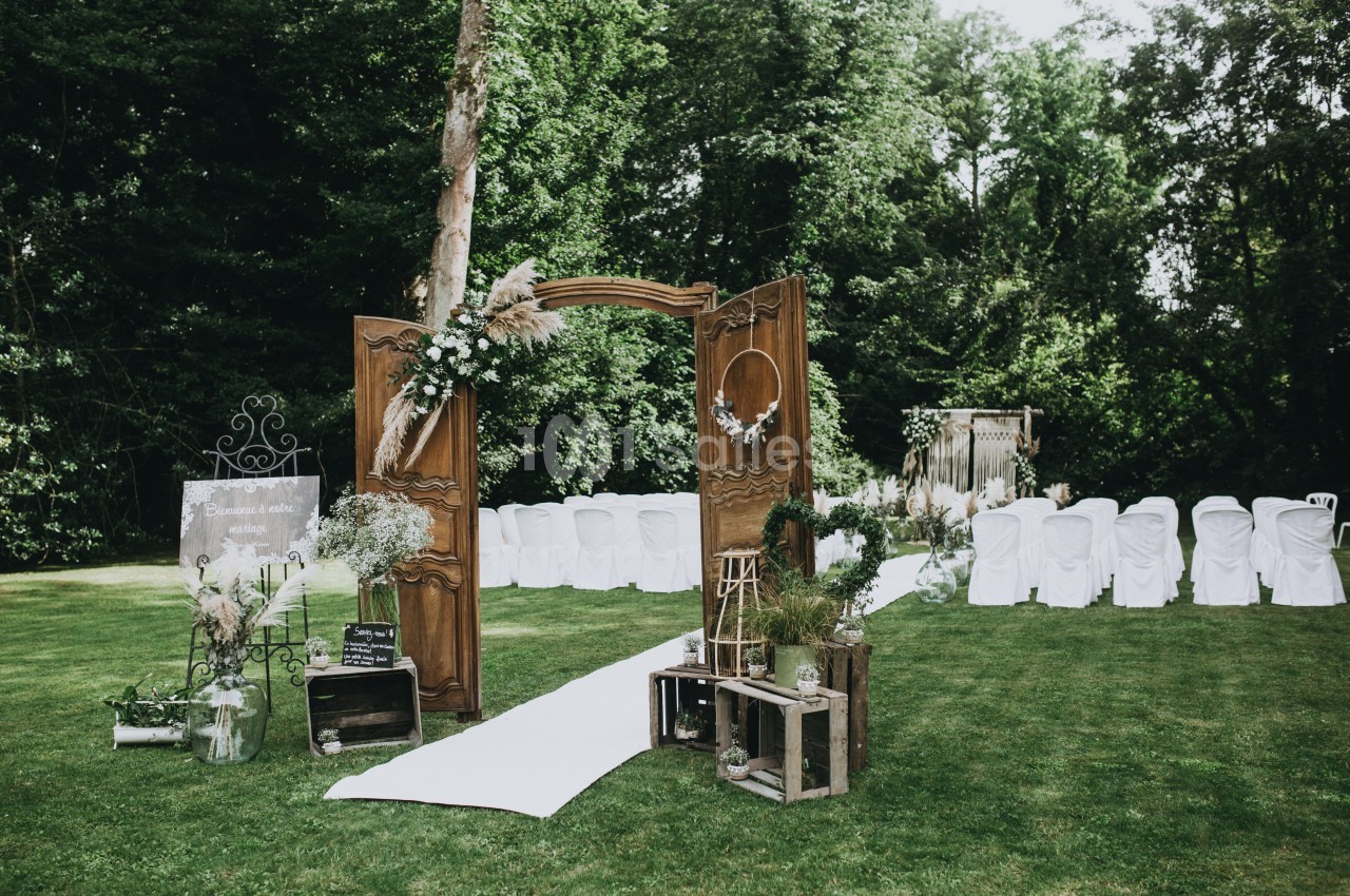 Allée de cérémonie en plein air avec chaises blanches, arche en bois décorée de fleurs et verdure environnante.