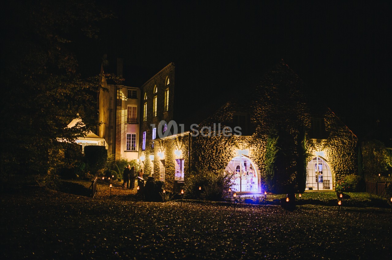 Bâtiments en pierre éclairés la nuit, avec des fenêtres illuminées et un jardin sombre au premier plan.