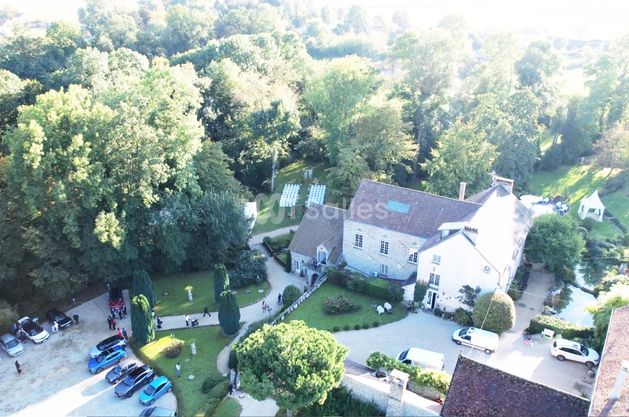 Vue aérienne d'une grande maison entourée de jardins, d'arbres et de voitures stationnées dans une cour.