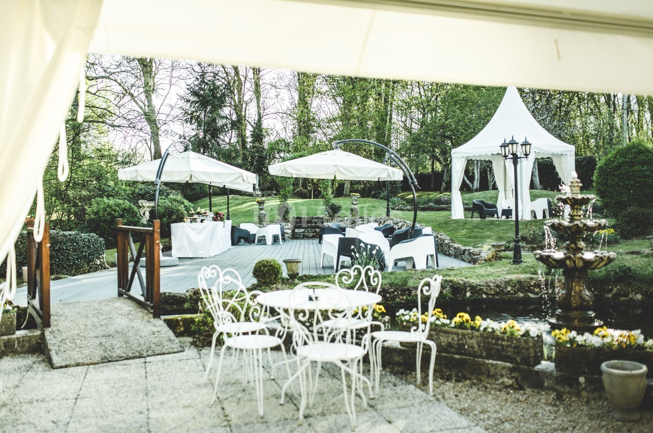 Jardin aménagé avec tables blanches, parasols, fontaine et tentes, entouré de verdure.