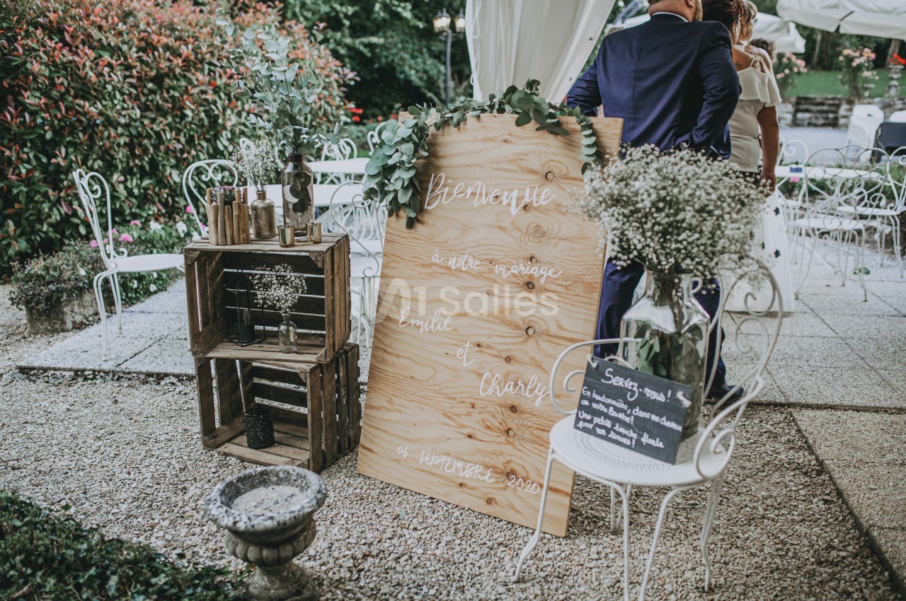 Panneau de bienvenue en bois décoré, entouré de fleurs et d'éléments rustiques, lors d'une cérémonie en extérieur.