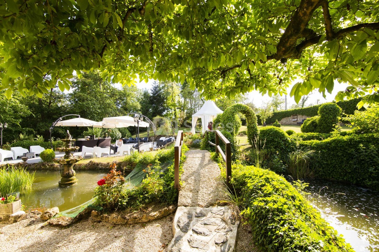 Jardin verdoyant avec un pont en bois, une fontaine, des parasols et un pavillon blanc sous un ciel ensoleillé.