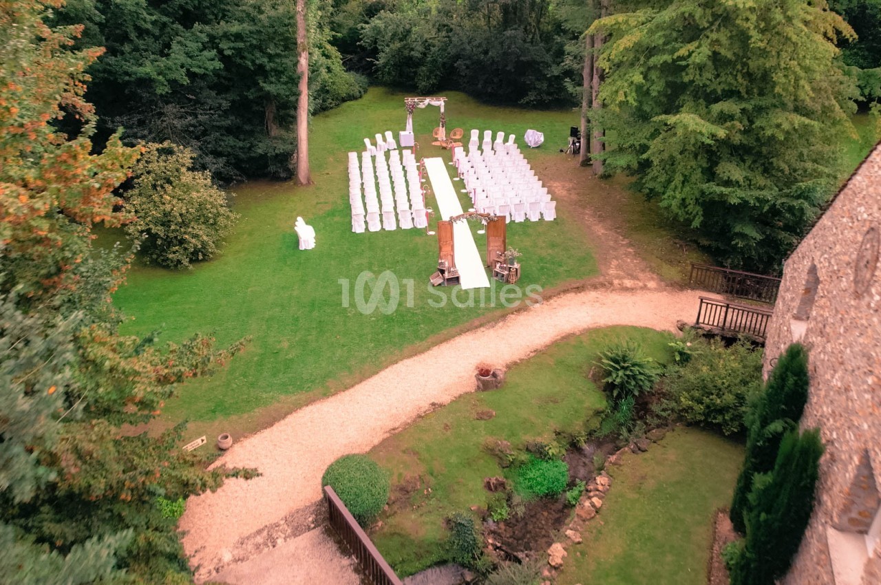 Vue aérienne d'une cérémonie de mariage en extérieur avec des chaises blanches alignées sur une pelouse entourée d'arbres.