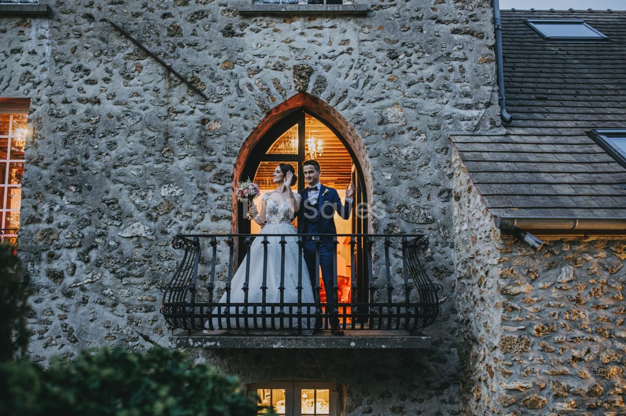 Un couple en tenue de mariage se tient sur un balcon en pierre devant une fenêtre illuminée.