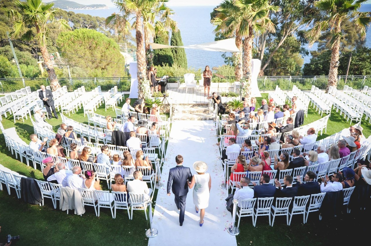 Un couple marche vers une cérémonie de mariage en plein air, entouré d'invités assis sur des chaises blanches.