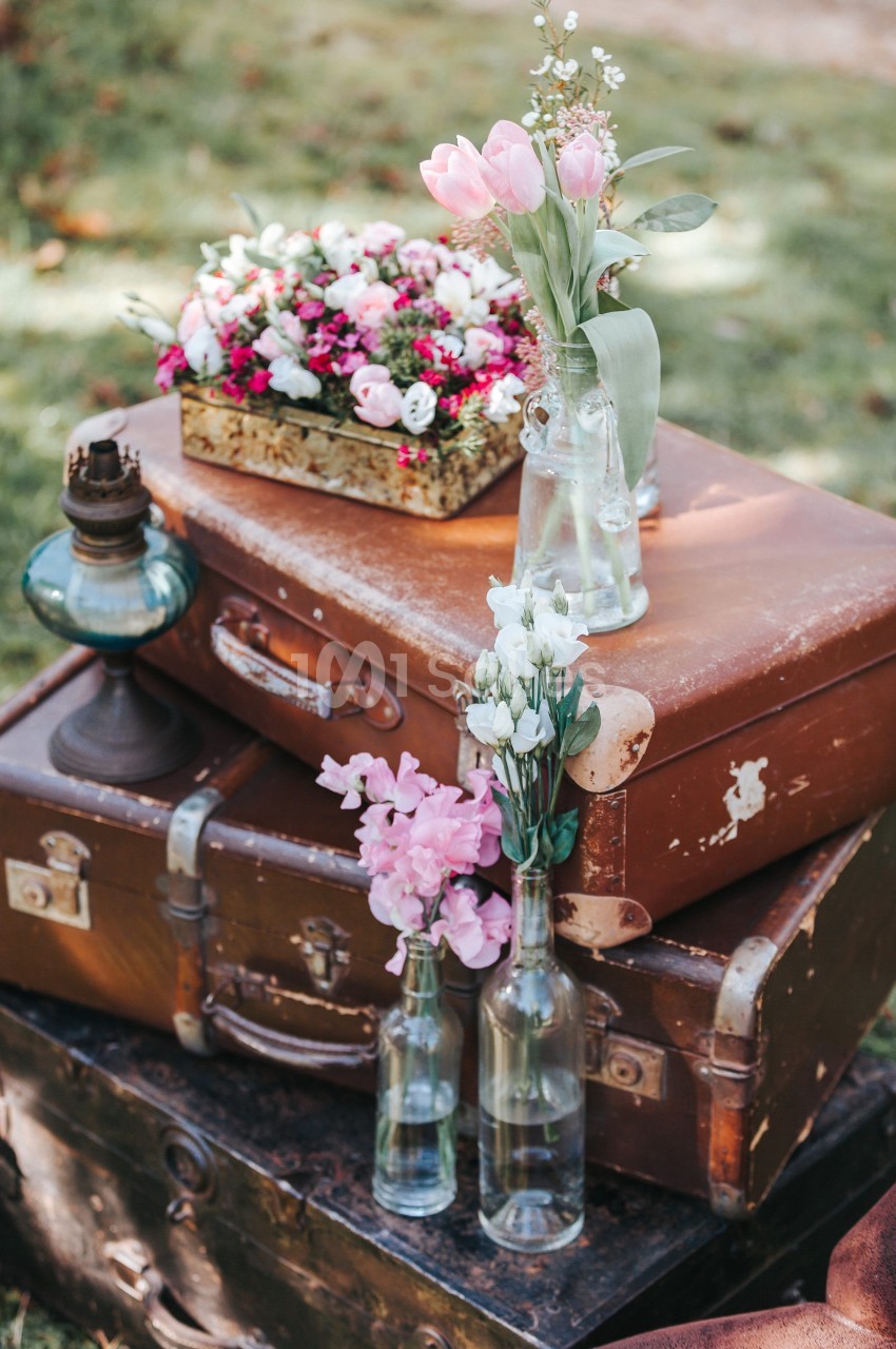 Des valises vintage empilées décorées de fleurs fraîches dans des vases en verre, disposées en extérieur.
