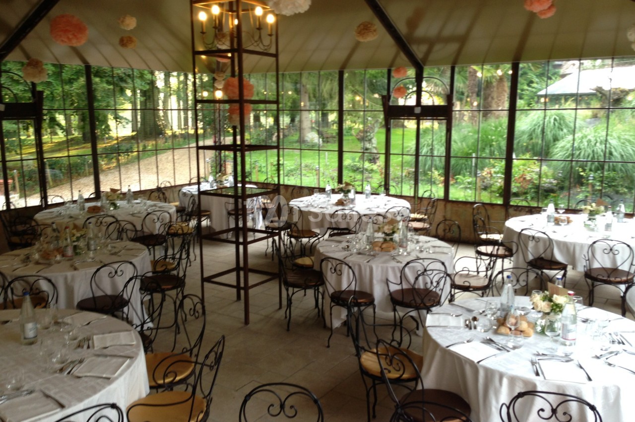 Salle de réception lumineuse avec des tables rondes dressées, entourée de grandes baies vitrées donnant sur un jardin…