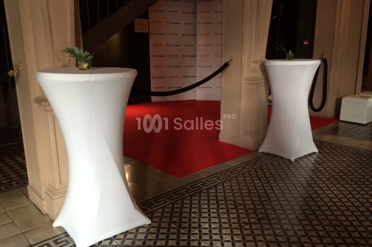 Deux tables hautes drapées de blanc encadrent un tapis rouge dans un espace intérieur au sol carrelé.