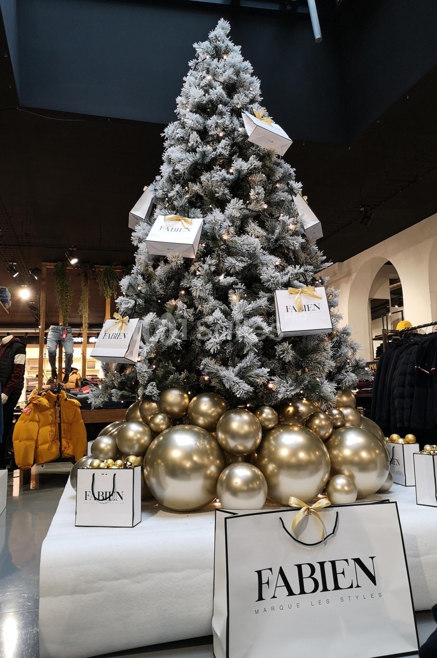 Sapin de Noël décoré de boules dorées et entouré de sacs cadeaux dans un magasin.