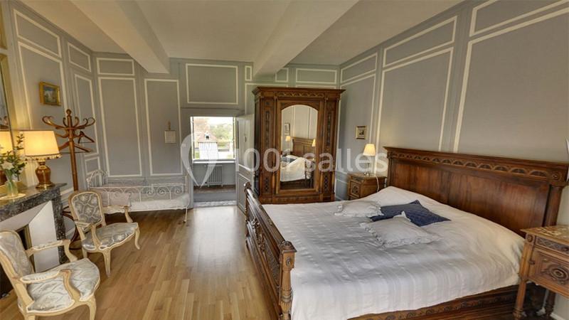 Chambre lumineuse avec lit double, armoire en bois sculpté, fauteuils et vue sur une fenêtre donnant sur l'extérieur.