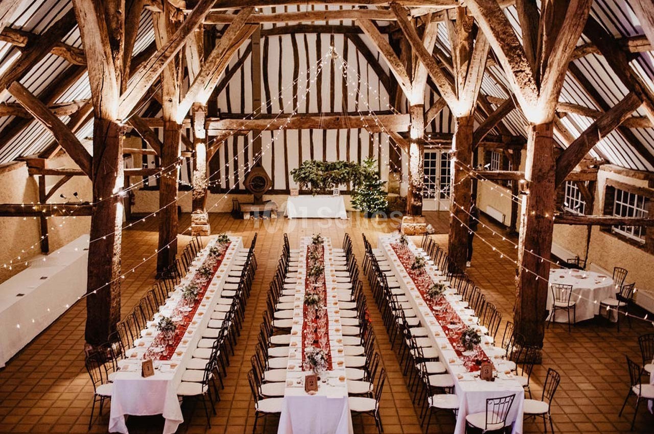 Grande salle de réception avec poutres apparentes, tables longues décorées de fleurs et guirlandes lumineuses suspendues.
