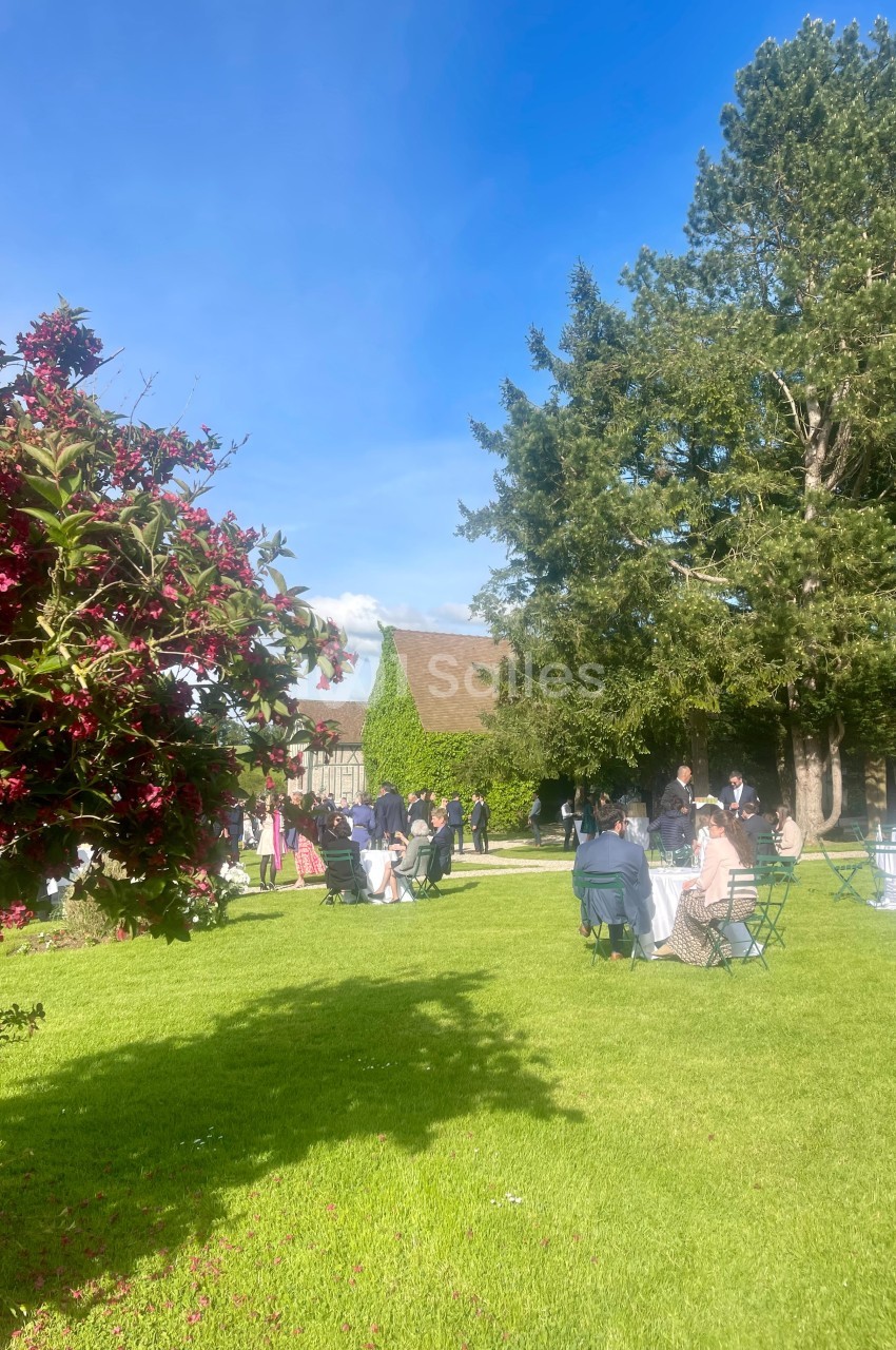 Personnes rassemblées dans un jardin verdoyant avec des tables, des arbres et un bâtiment en arrière-plan par temps…