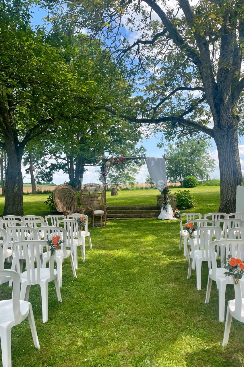 Chaises blanches alignées en extérieur face à une arche décorée, sous des arbres dans un cadre verdoyant.