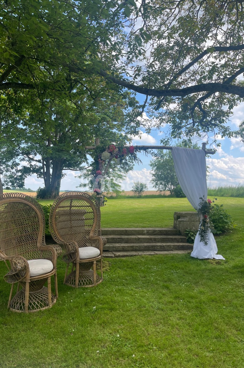 Deux fauteuils en rotin et une arche décorée de fleurs et de tissu blanc dans un jardin verdoyant.