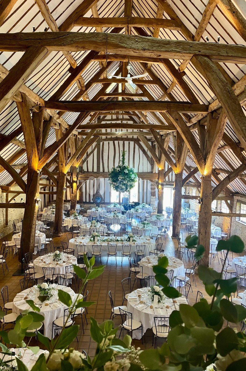 Grande salle de réception avec charpente en bois apparente, tables rondes dressées et décoration florale suspendue.