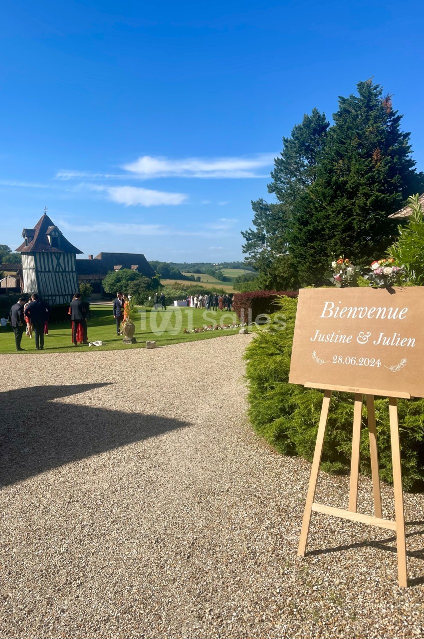 Panneau de bienvenue pour un mariage, placé sur un chevalet dans un jardin avec des invités en arrière-plan.