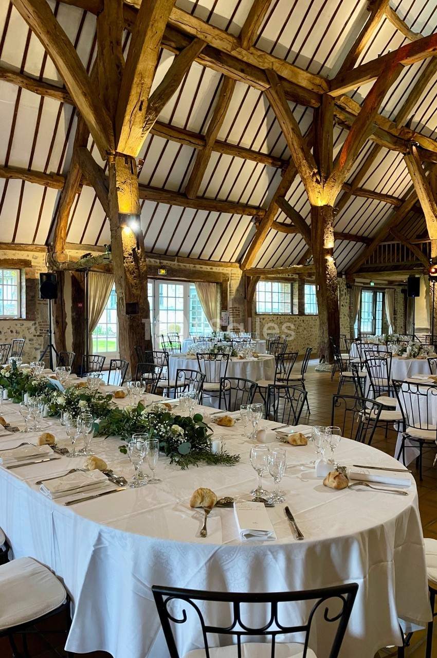 Salle de réception avec charpente en bois, tables dressées avec nappes blanches, vaisselle élégante et décoration florale.