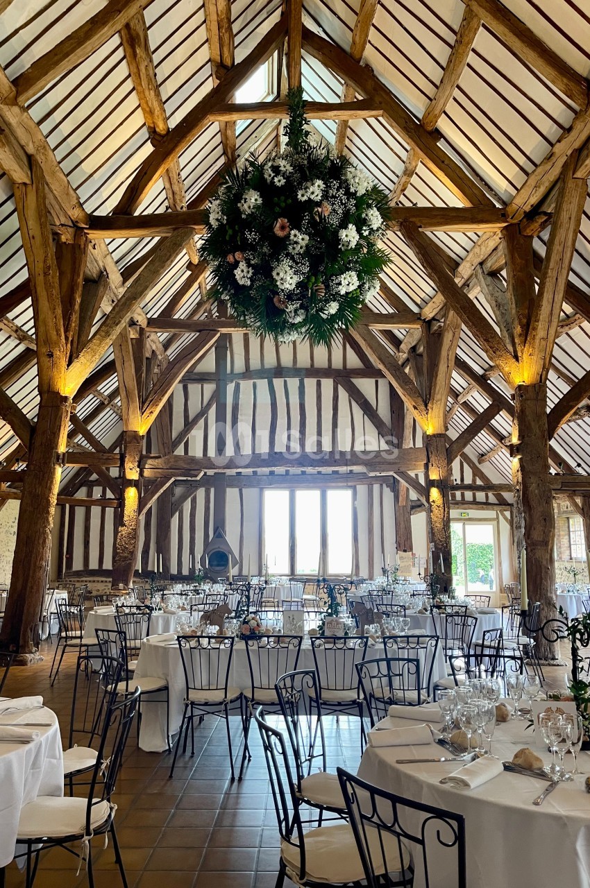 Salle de réception rustique avec poutres apparentes, tables dressées et une grande décoration florale suspendue.