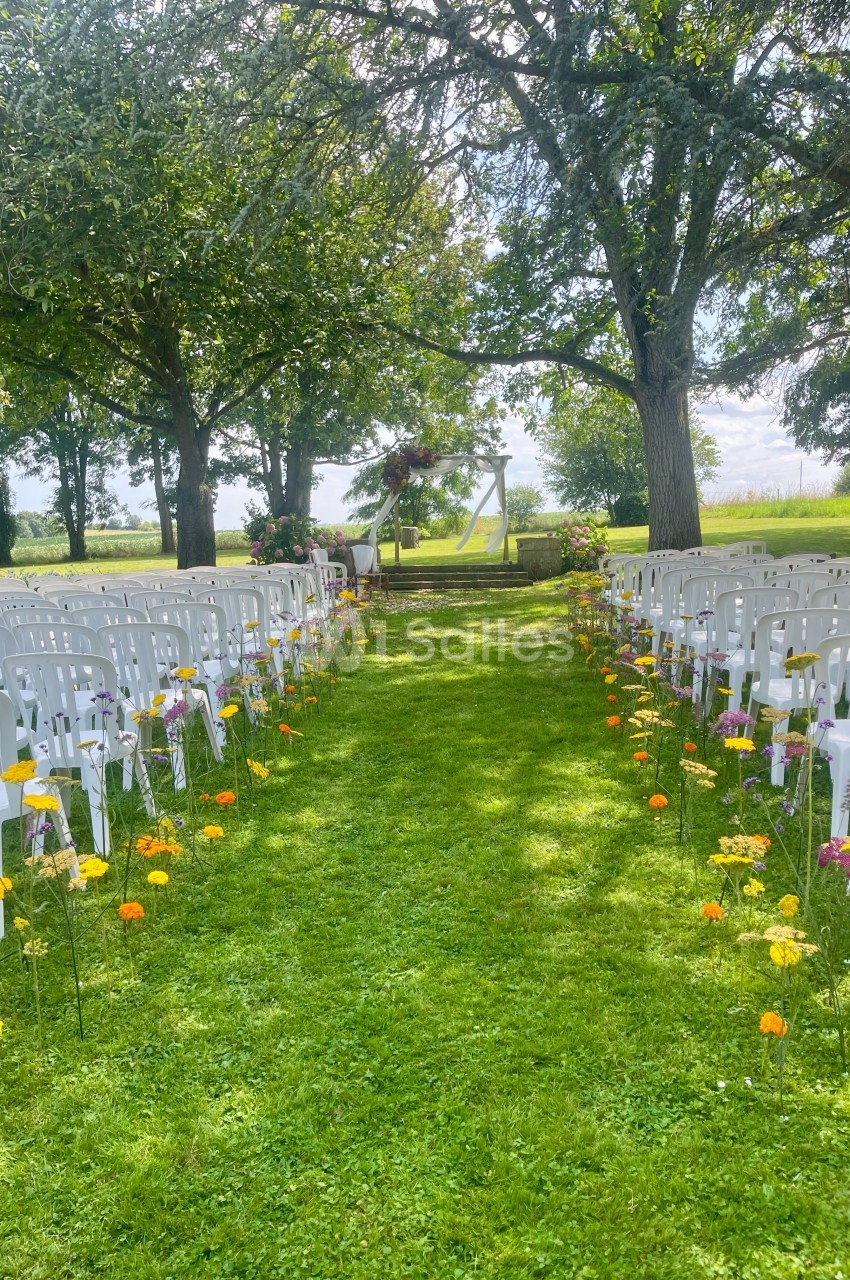 Allée centrale bordée de fleurs menant à une arche décorée pour une cérémonie en plein air, entourée d'arbres.