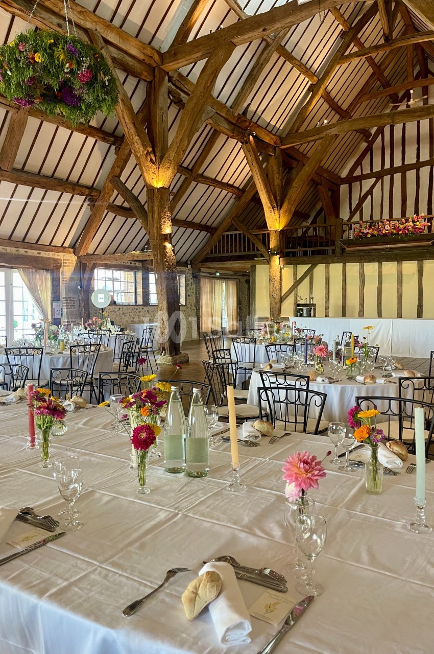 Salle de réception rustique avec poutres apparentes, tables dressées, décor floral coloré et lumière naturelle.