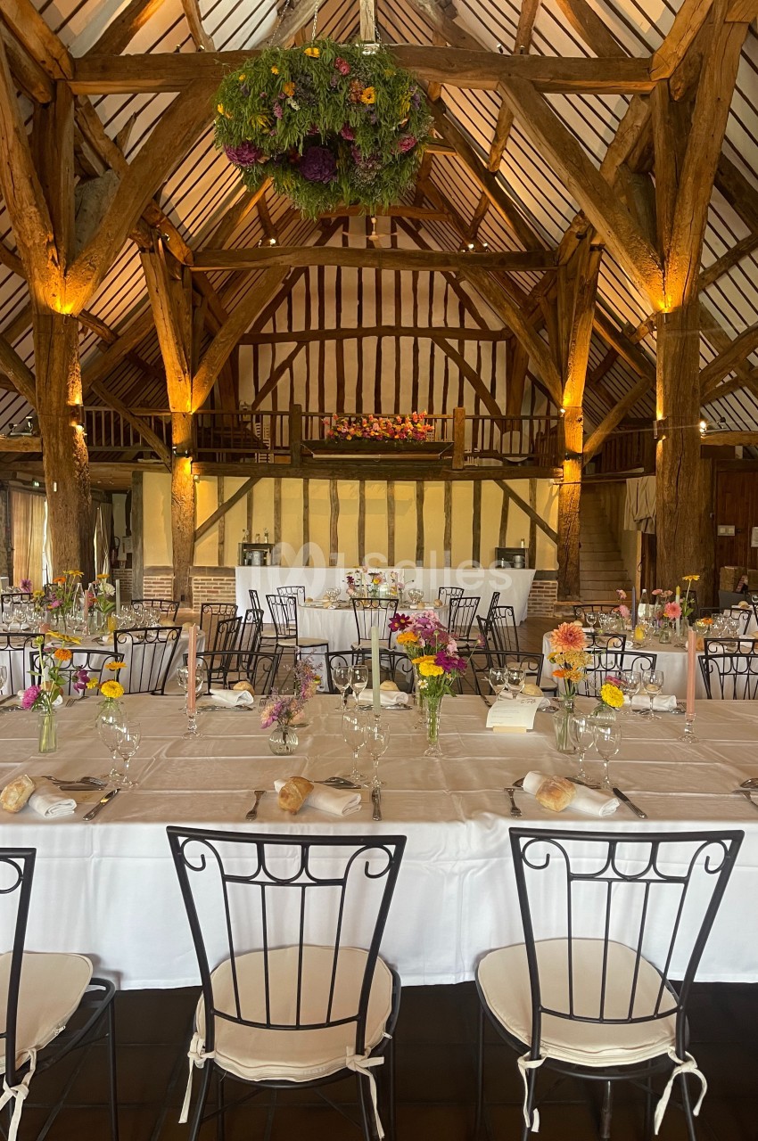Salle de réception rustique avec poutres apparentes, tables dressées et décorations florales colorées.