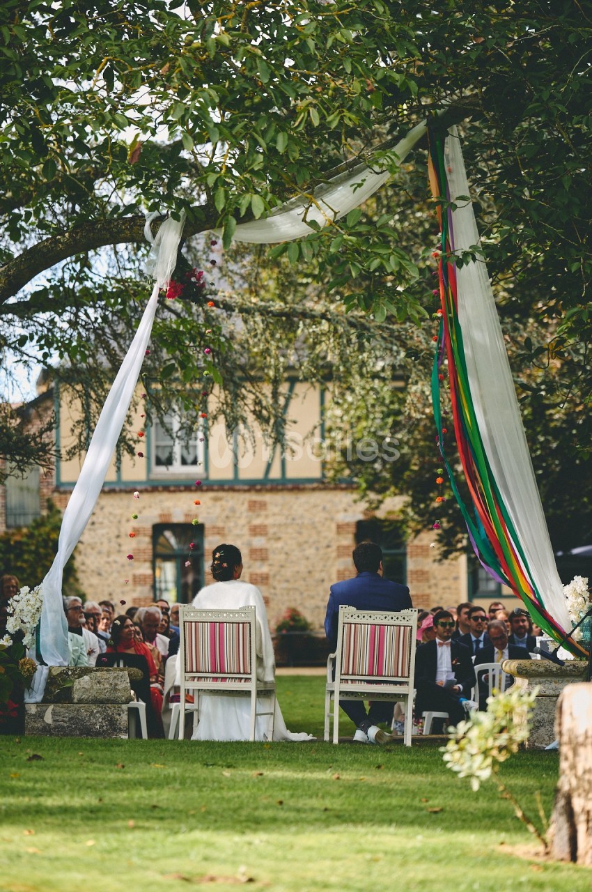 Un couple assis sous un arbre décoré, face à des invités lors d'une cérémonie en plein air.