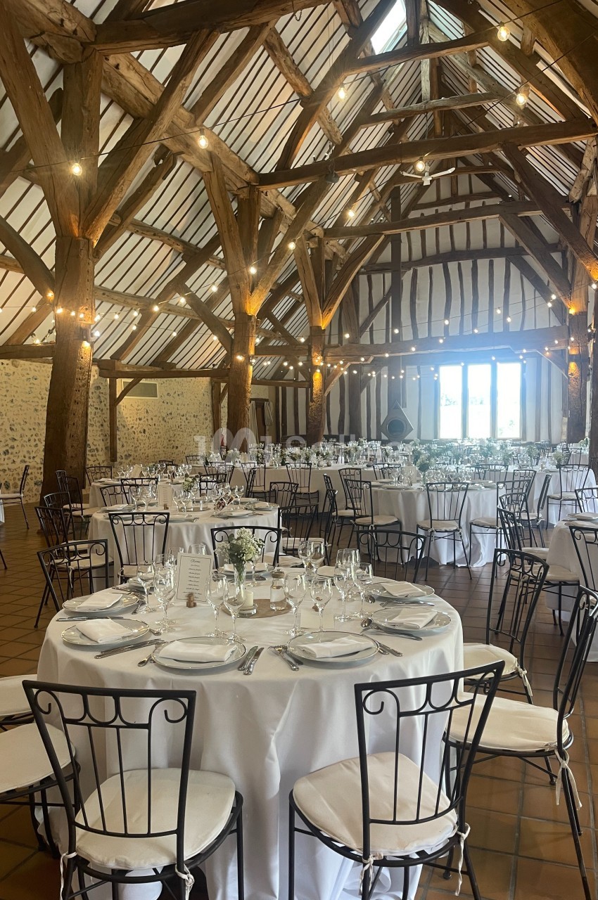 Salle de réception avec charpente en bois apparente, tables rondes dressées avec nappes blanches et chaises en métal.