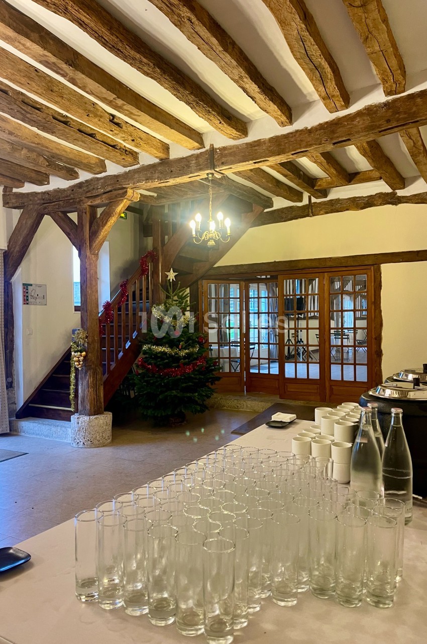 Salle avec poutres apparentes, table dressée avec verres et carafes, escalier en bois et sapin décoré en arrière-plan.