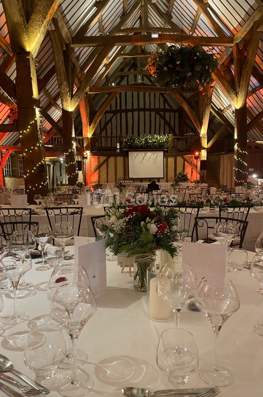 Grande salle de réception avec charpente apparente, tables dressées, décorations florales et éclairage chaleureux.