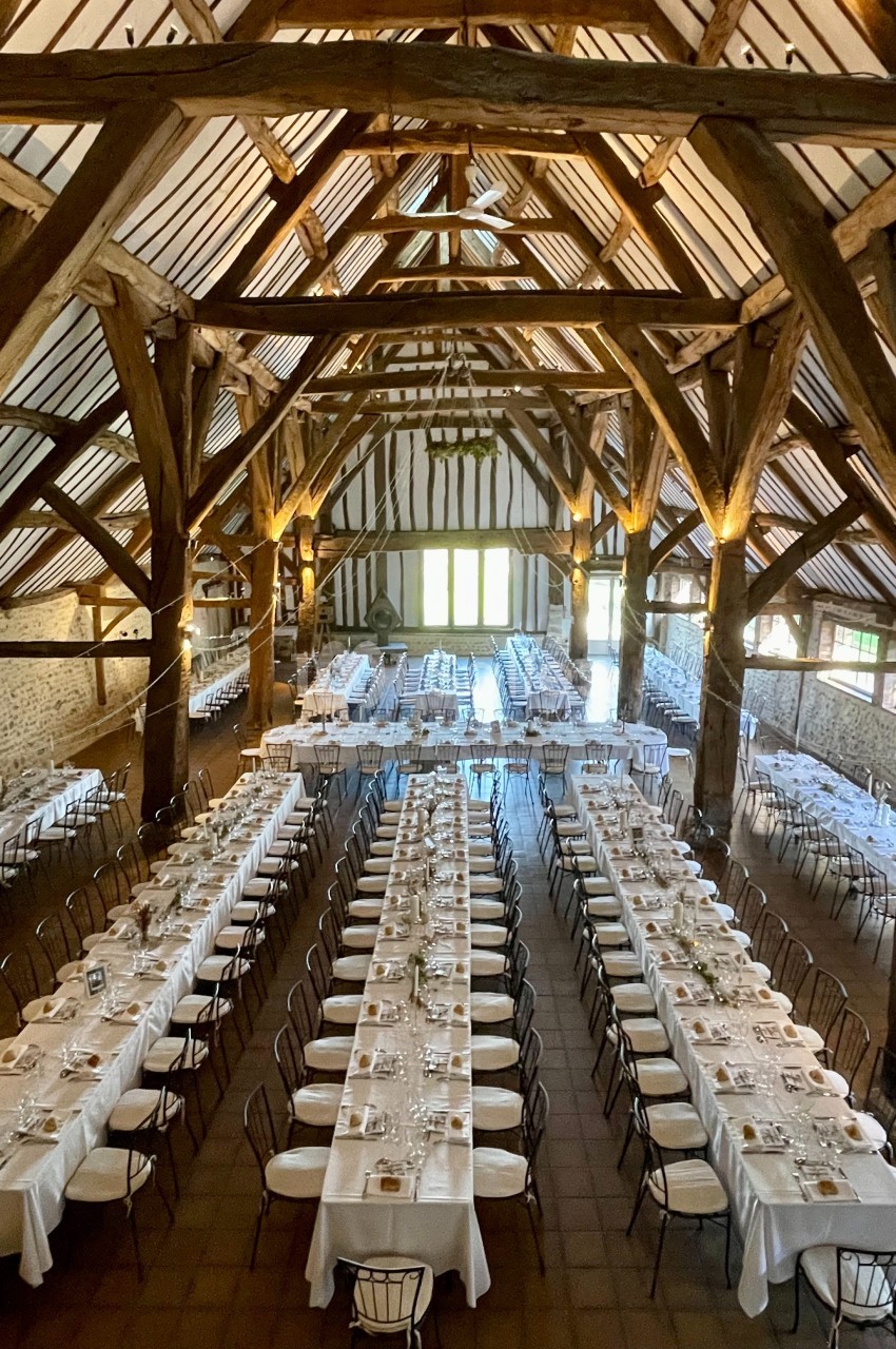 Salle de réception rustique avec de longues tables dressées pour un événement, sous une charpente en bois apparente.