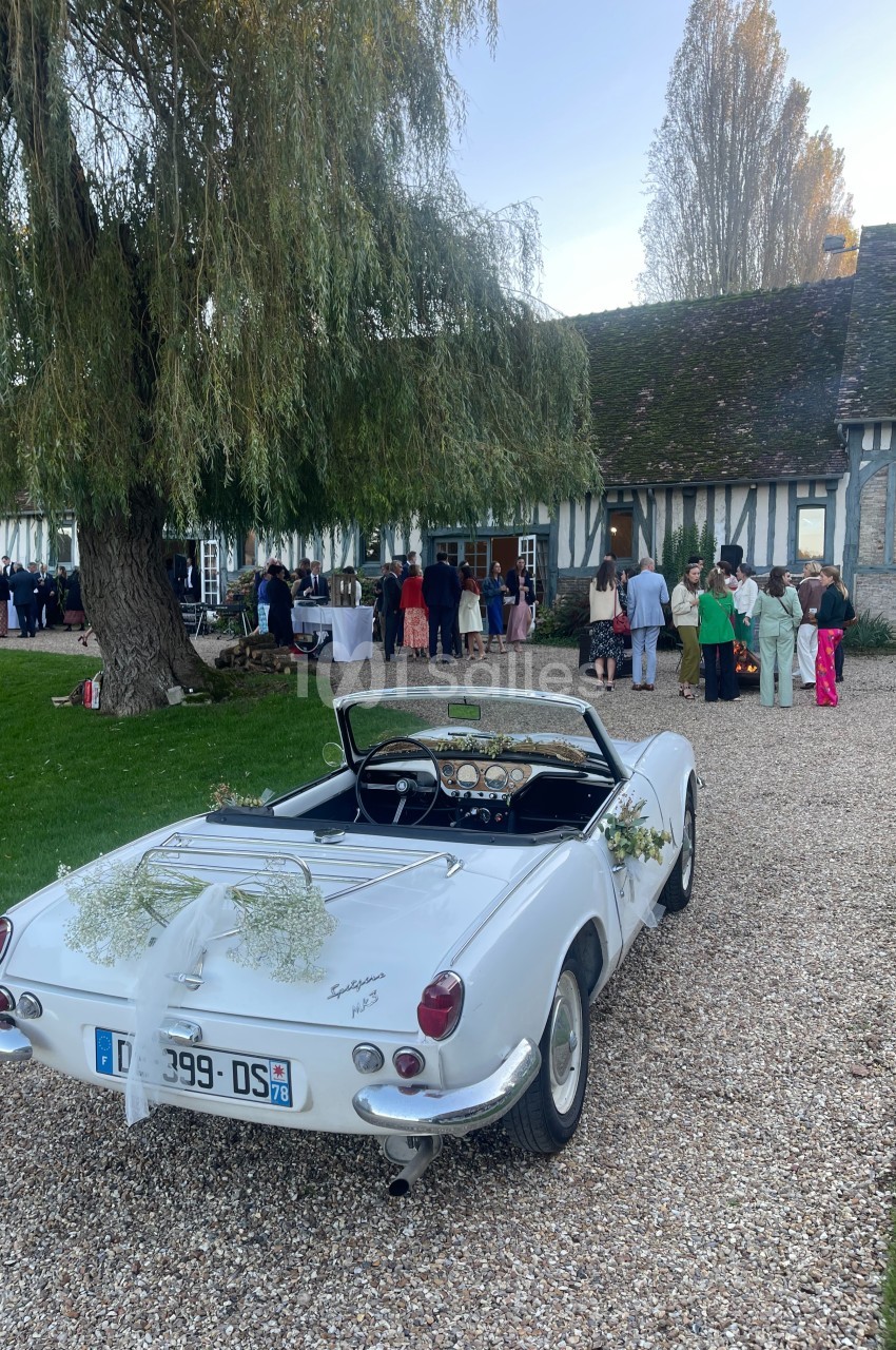 Voiture décapotable blanche décorée pour un mariage, stationnée devant une maison à colombages avec des invités à l'arrière…