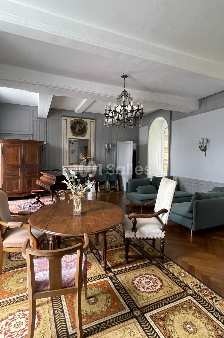 Salle de séjour lumineuse avec mobilier ancien, tapis coloré, table en bois, fauteuils et lustre suspendu.